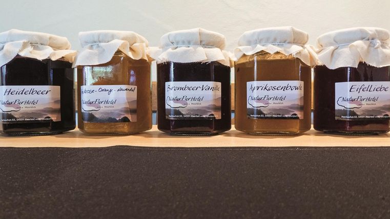 Eine Reihe von fünf Marmeladengläsern auf einem Tisch. Jedes Glas hat ein Etikett mit der Geschmacksrichtung, darunter Heidelbeer, Erdbeere und mehrere andere.