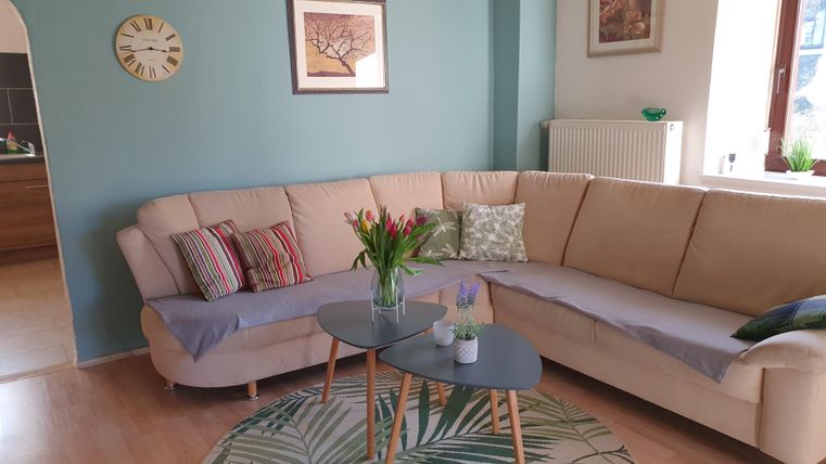 Ein gemütliches Wohnzimmer mit einer beigen Couch und einem modernen Tisch. An der Wand hängen Bilder, und frische Blumen schmücken den Tisch.