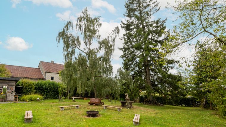 Eine grüne Wiese mit Holzbänken und einem Feuerplatz in der Mitte. Im Hintergrund stehen große Bäume und ein Haus.