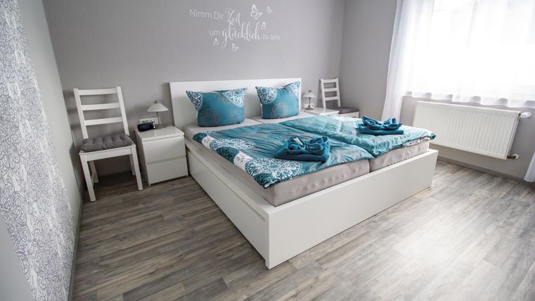Ein gemütliches Schlafzimmer mit einem großen Bett und blauen Kissen. Der Raum ist hell und modern eingerichtet, mit Holzfußboden und viel Tageslicht.