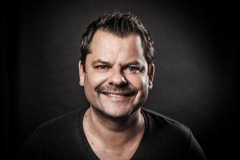 Ein lächelnder Mann mit kurzem Haar und Schnurrbart. Er trägt ein schwarzes T-Shirt und der Hintergrund ist dunkel.