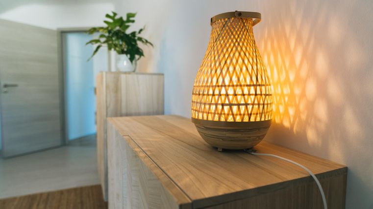 Eine stilvolle Lampe auf einem Holzschrank, die warmes Licht ausstrahlt. Im Hintergrund ist eine Pflanze und eine offene Tür zu sehen.