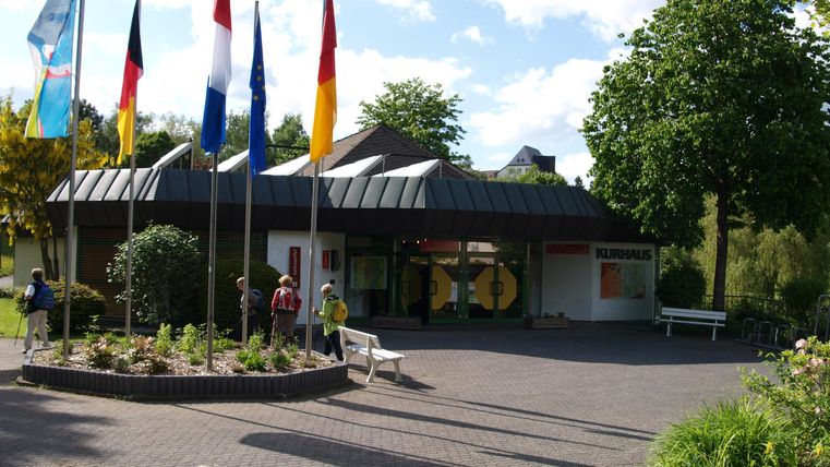Ein Eingangsbereich mit mehreren Fahnen und einem modernen Gebäude. Bäume und Pflanzen umgeben die Szenerie, und der Himmel ist teilweise bewölkt.