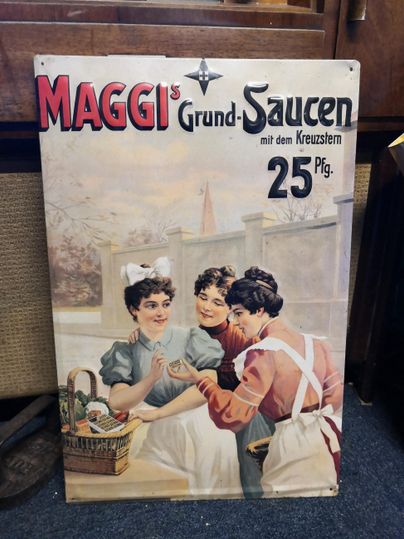 Ein altes Werbeschild für Maggi-Saucen, das drei Frauen in historischen Kleidern zeigt. Die Frauen stehen in einer Küche und interagieren miteinander, während im Hintergrund ein Garten zu sehen ist.