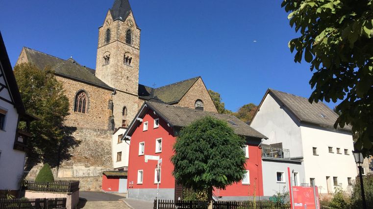 Eine malerische Straße mit alten Häusern und einer beeindruckenden Kirche im Hintergrund. Der Himmel ist klar und blau, was die Szene freundlich macht.
