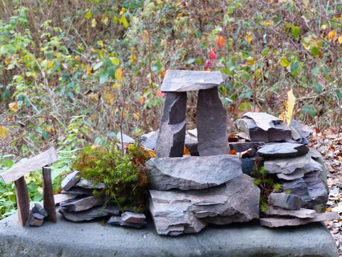 Eine kleine Steinskulptur, die an eine prähistorische Megalithstruktur erinnert. Umgeben von Natur mit buntem Laub im Hintergrund.