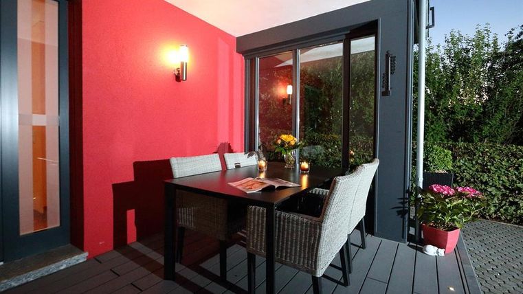 Eine gemütliche Terrasse mit einem Tisch und Stühlen. Im Hintergrund sieht man Pflanzen und eine rot gestrichene Wand.