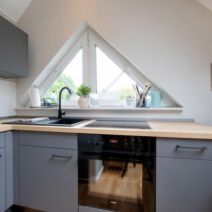 Eine moderne Küche mit grauen Schränken und einer Holzarbeitsplatte. Große Fenster sorgen für viel natürliches Licht und eine angenehme Atmosphäre.