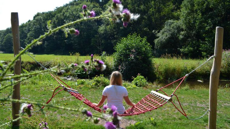 Person sitzt in einer roten Hängematte in einem grünen Park, umgeben von Bäumen und Blumen.
