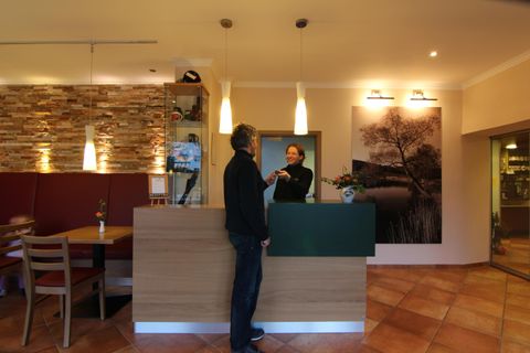 Ein Empfangsbereich in einem Hotel mit einem freundlichen Mitarbeiter und einem Gast. Die Atmosphäre ist gemütlich und einladend, mit warmen Farben und moderner Einrichtung.