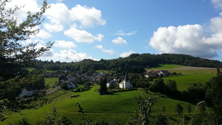 Eine malerische Landschaft mit einem kleinen Dorf und einer Kirche. Der Himmel ist blau mit einigen Wolken und die Umgebung ist grün und hügelig.