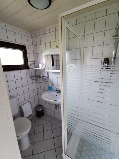 Ein modernes Badezimmer mit einer Dusche, einem Waschbecken und einer Toilette. Die Wände sind mit weißen Fliesen gefliest und es gibt ein Fenster für natürliches Licht.
