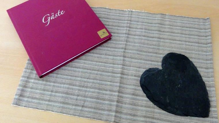 Ein rotes Gästebuch liegt auf einem gestreiften Tuch. Neben dem Buch befindet sich ein schwarzes Herz aus Stein.
