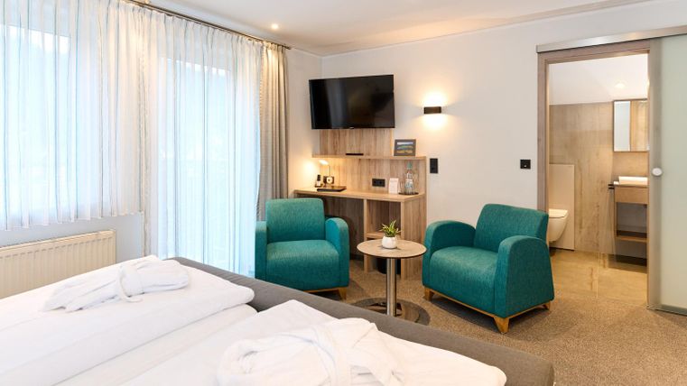 Ein modernes Hotelzimmer mit zwei Betten, einer Sitzecke und einem Fernseher. Die hellen Vorhänge und die grünen Sessel sorgen für eine freundliche Atmosphäre.
