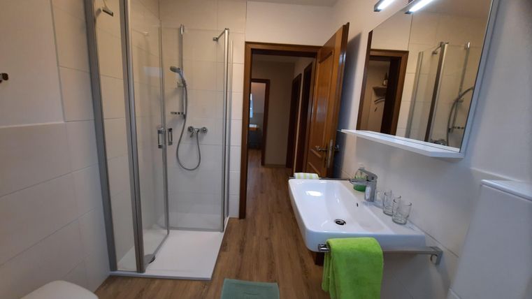 Ein modernes Badezimmer mit einer Dusche und einem Waschbecken. Die Wände sind hell gefliest und es gibt Holzdetails im Boden.