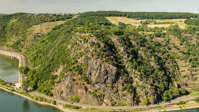 A picturesque landscape with rolling hills and wooded areas. In the foreground, a river flows along the riverside road.