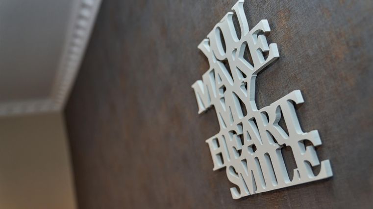 Ein dekoratives Wandbild mit der Aufschrift „You make my heart smile“. Es steht auf einer neutralen Wandfarbe.