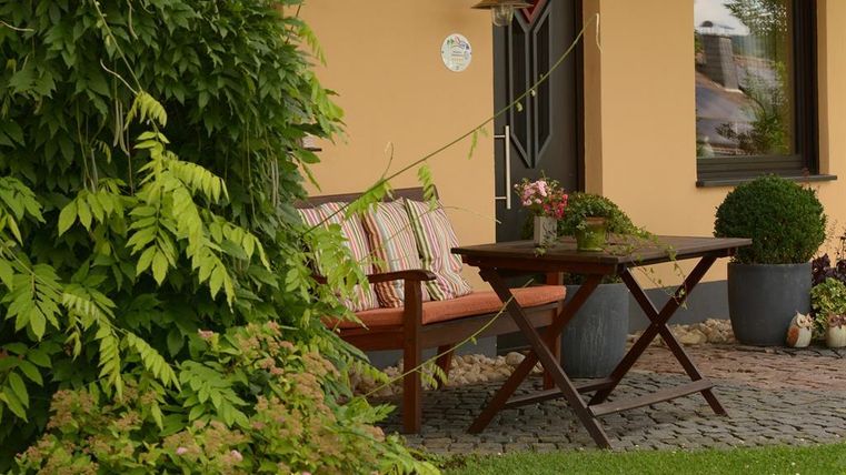Ein gemütlicher Außenbereich mit einer Bank und einem Tisch. Umgeben von grünen Pflanzen und einem einladenden Ambiente.