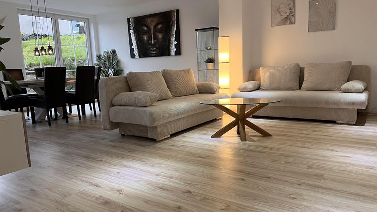 Ein modernes Wohnzimmer mit zwei beigen Sofas und einem Holztisch. An der Wand hängt ein Kunstwerk und im Hintergrund sind ein Essbereich und Fenster mit Blick ins Grüne sichtbar.