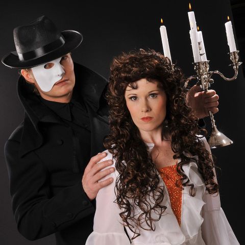 Ein Mann in einem schwarzen Mantel und einer Maske umarmt eine Frau mit lockigem Haar, die ein elegantes Kleid trägt. In der Hand des Mannes befindet sich ein Kerzenständer mit brennenden Kerzen.