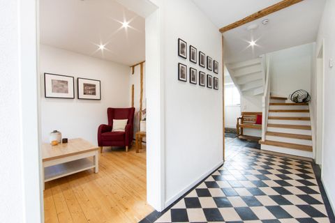 A bright hallway with a checkered floor and a staircase. On the left side, there is a red armchair and some pictures on the wall.