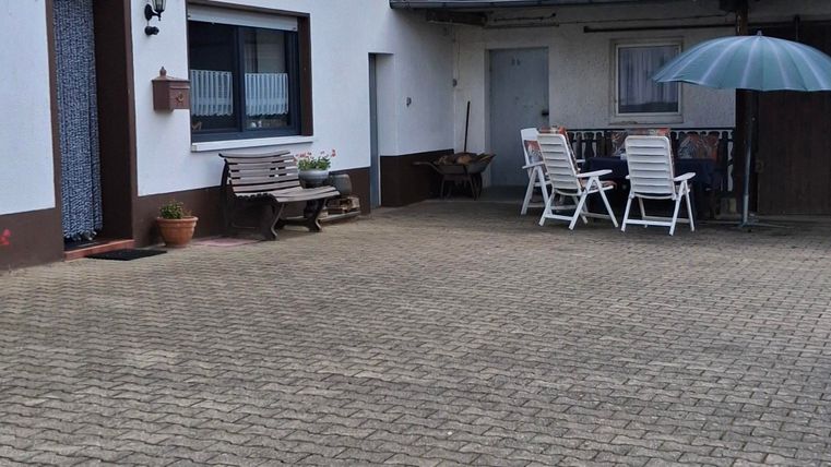 Ein ruhiger Innenhof mit einem weißem Haus und einem überdachten Tisch. Im Hintergrund ist die ausenterrasse mit Tisch und Stühlen