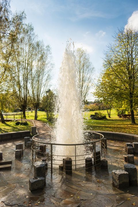 Ein schöner Springbrunnen in einem Park, umgeben von Bäumen. Das Wasser spritzt hoch in den klaren Himmel.