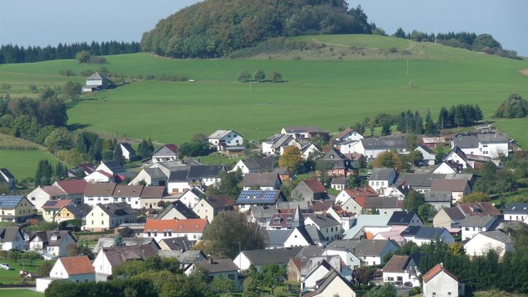 Eine malerische Landschaft mit einem kleinen Dorf, umgeben von grünen Wiesen und Hügeln. Die Häuser sind bunt und gut angeordnet, was eine idyllische Atmosphäre schafft.
