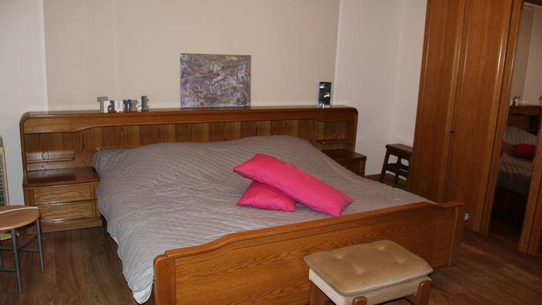 Ein gemütliches Schlafzimmer mit einem großen Holzbett und violetten Kissen. Die Wände sind in neutralen Farben gehalten, und es gibt einige Möbelstücke aus Holz.
