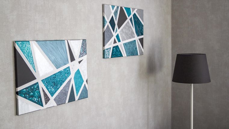 Zwei moderne Wandbilder mit geometrischen Mustern in Blau- und Grautönen. Eine elegante Stehlampe steht daneben und ergänzt die Einrichtung.
