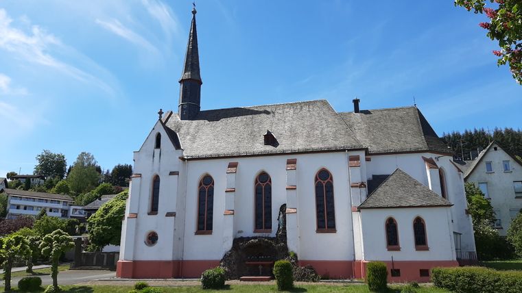 Weiße Kirche mit spitzem Turm und roten Fenstern in Deudesfeld, umgeben von Bäumen und blauen Himmel.