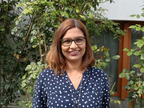Eine lächelnde Frau mit braunen Haaren steht vor einer grünen Hecke. Sie trägt ein gepunktetes Oberteil und eine Brille.