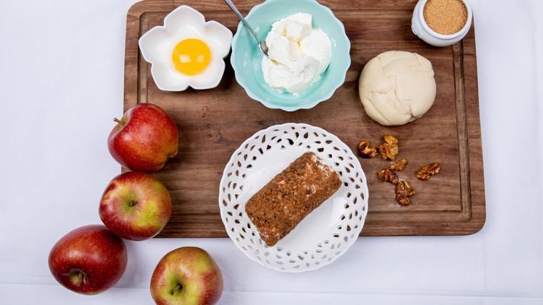 Ein Holzbrett mit frischen Äpfeln, einem Stück Kuchen und einer Schüssel mit Sahne. Zudem sind ein rohes Ei, ein Stück Teig und ein Glas Zucker zu sehen.