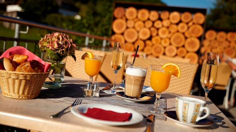 Ein Tisch im Freien mit einer Auswahl an Getränken, darunter Orangensaft und Kaffee, sowie einer kleinen Korb mit Gebäck. Im Hintergrund sind Holzscheite zu sehen, die eine gemütliche Atmosphäre schaffen.