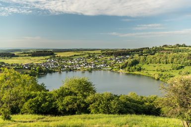 Eine malerische Landschaft mit einem klaren See und sanften Hügeln. Im Hintergrund sieht man ein kleines Dorf umgeben von Bäumen.