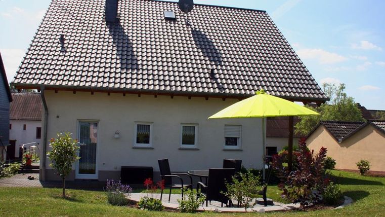 Ein modernes Haus mit einem Steildach und einer sonnigen Terrasse. Ein gelber Sonnenschirm bietet Schatten auf dem Rasen.
