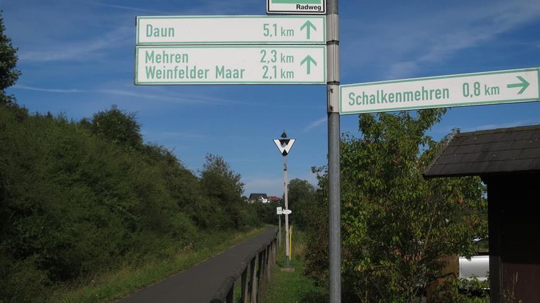 Ein Wegweiser zeigt die Richtungen zu verschiedenen Orten: Daun, Mehrenh, Weinfelder Maar und Schalkenmehrn. Die Umgebung ist grün und friedlich, ideal für Radfahrer.