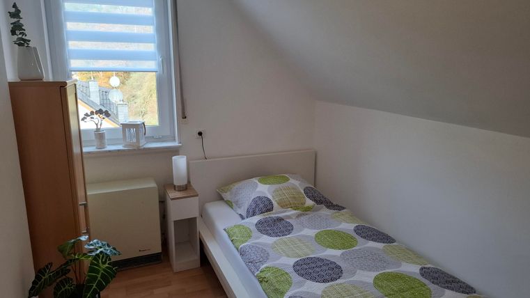 Ein kleines, helles Schlafzimmer mit einem Einzelbett und bunten Kissen. Neben dem Bett steht ein Nachttisch und ein Regal, und ein Fenster sorgt für Tageslicht.