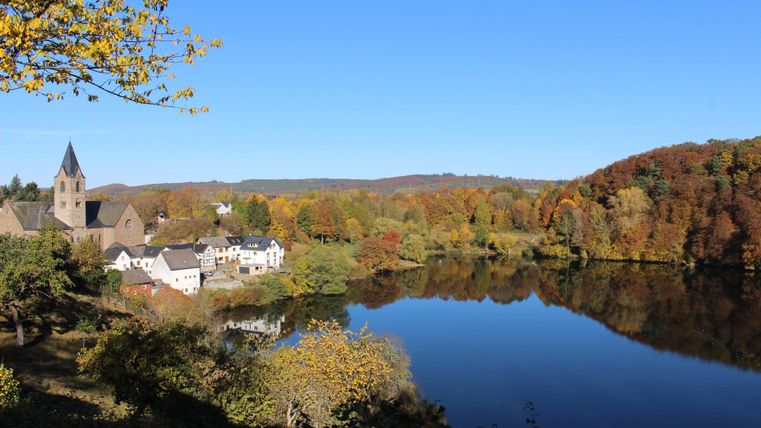 Eine idyllische Landschaft mit bunten Herbstbäumen und einem ruhigen See. Im Hintergrund ist eine Kirche und ein kleines Dorf zu sehen.