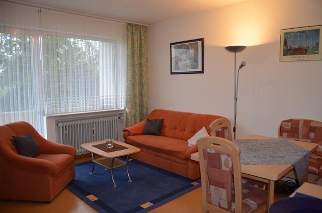 Ein gemütliches Wohnzimmer mit zwei orangenen Sofas und einem Tisch. Die Fenster sind mit Vorhängen bedeckt und die Wände sind mit Bildern dekoriert.