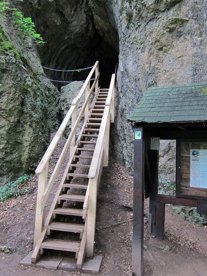Eine Holztreppe führt zu einem Höhleneingang. Daneben steht eine Infotafel mit einem kleinen Dach. Der Eingang ist von Felsen umgeben.