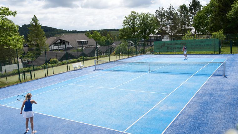 Ein Tennisplatz mit blauen Belag und zwei Spielerinnen, die ein Match spielen. Im Hintergrund sind Bäume und Häuser zu sehen.