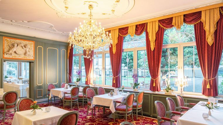 Ein elegantes Restaurant mit dekorativen roten Vorhängen und einem großen Kronleuchter. Die Tische sind geschmackvoll gedeckt und bieten einen Blick auf den Außenbereich.