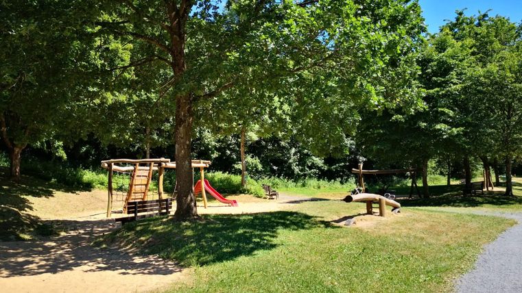 Ein Spielplatz mit einer Rutsche und einem Klettergerüst unter großen Bäumen. Die Umgebung ist grün und einladend.