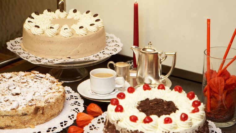 Drei verschiedene Torten stehen auf einem Tisch, umgeben von kleinen dekorativen Details. Dazu gibt es eine Kaffeetasse, eine Teekanne und frische Früchte.