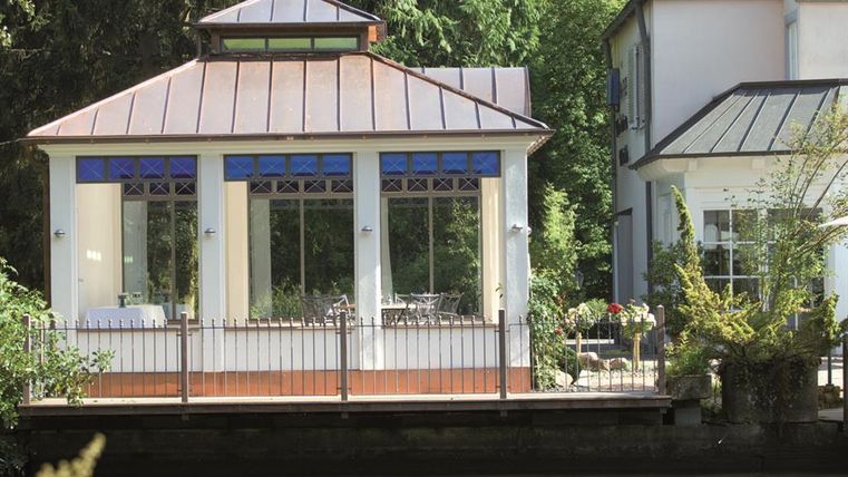 Ein gepflegtes Gartenhaus mit großen Fenstern und einem überdachten Bereich. Es liegt an einem ruhigen Gewässer, umgeben von Pflanzen und Bäumen.