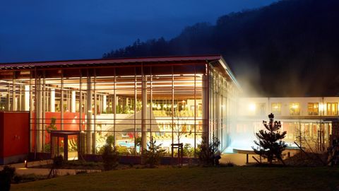 Ein modernes Gebäude mit großen Fenstern und rotem Dach bei Nacht. Im Hintergrund sind Berge und dampfendes Wasser zu sehen.