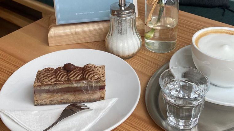 Ein Stück Torte auf einem Teller, daneben ein Glas Wasser und eine Tasse Kaffee. Im Hintergrund sind ein Zuckergefäß und eine Flasche mit Wasser zu sehen.