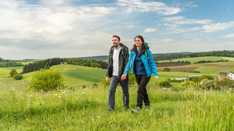 Een stel wandelt door een groen landschap met heuvels en een blauwe lucht.