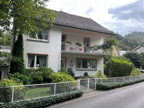 Ein zweigeschossiges, weißes Haus mit einem Dach und Balkon. Umgeben von einem gepflegten Garten und einer Hecke.
